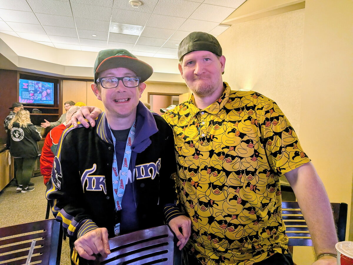 Two friends smiling arm in arm in the suite, one in a Vikings jacket and Twins lanyard, the other in a rubber duck shirt