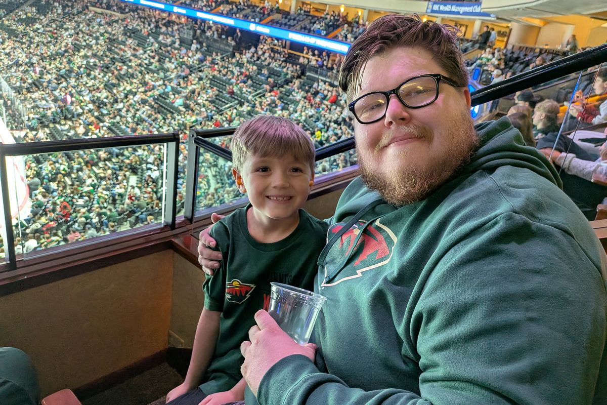 A man and a young boy smiling in the suite with the arena crowd behind them